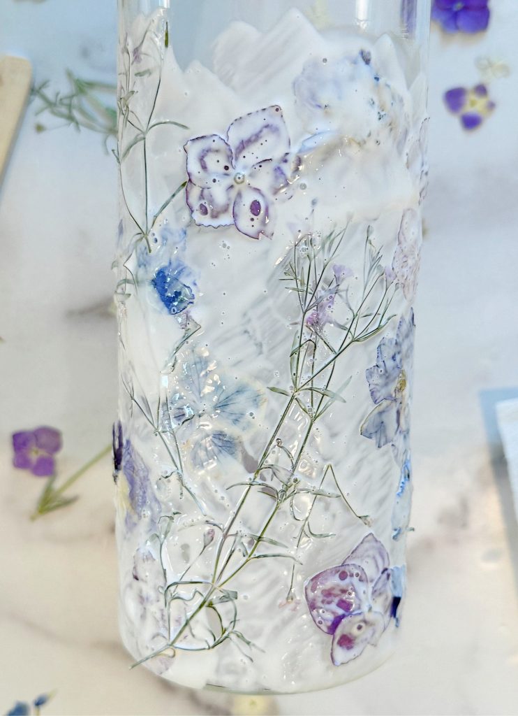 A close-up of a clear cylindrical vase adorned with pressed purple and white flowers and delicate green stems. The background is blurred, showing scattered flower petals on a light surface.