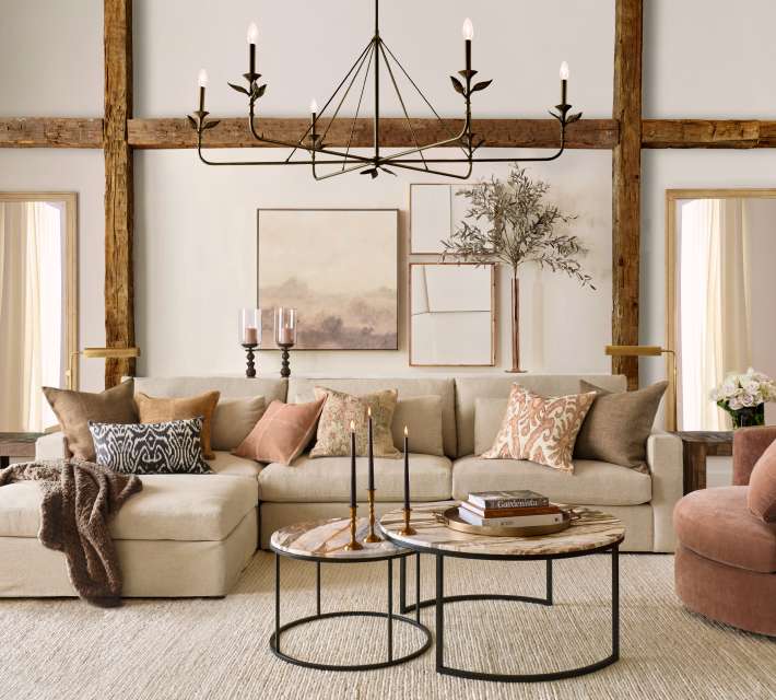 A cozy living room featuring a beige sectional sofa with assorted pillows, a round coffee table with candles, wall art, and a large chandelier. The walls have wooden beams and two framed mirrors.