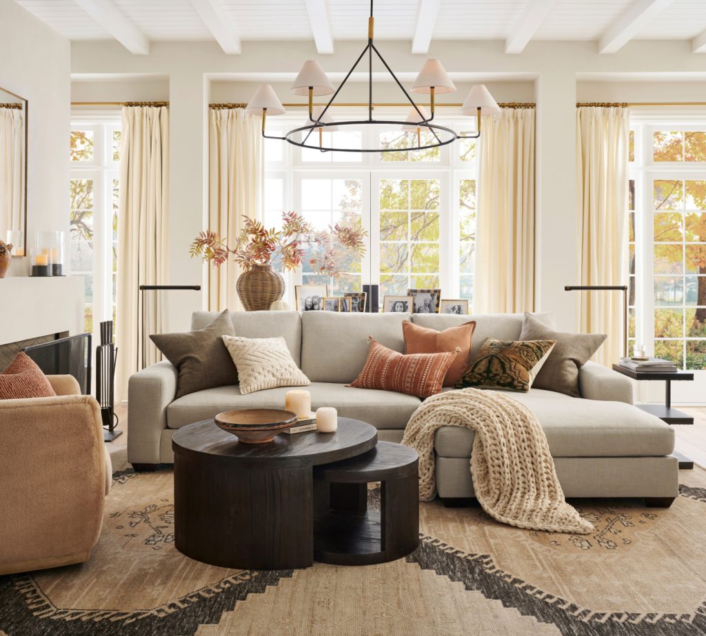 A modern living room with a beige sectional sofa, round nested coffee tables, a large rug, decorative throw pillows, and a knit blanket. Windows with light curtains let in natural light.