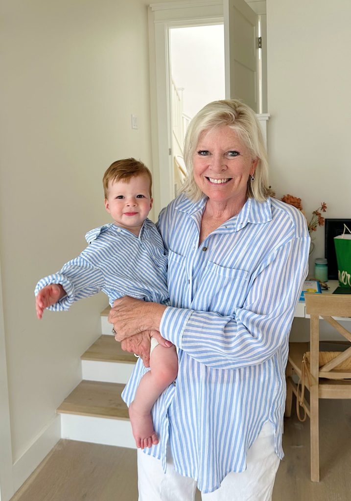 A woman wearing a blue and white striped shirt holds a smiling baby, also in a blue and white striped shirt, standing in a bright room with a staircase and white walls in the background.