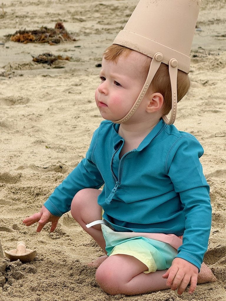 A young child sits on the sand at a beach, wearing a blue shirt and green shorts, with a sand bucket on their head.