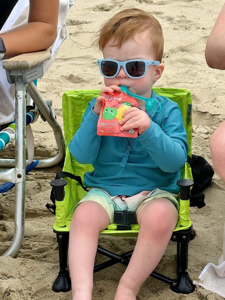 A young child wearing sunglasses sits in a small beach chair, drinking from a pouch while on the sand.