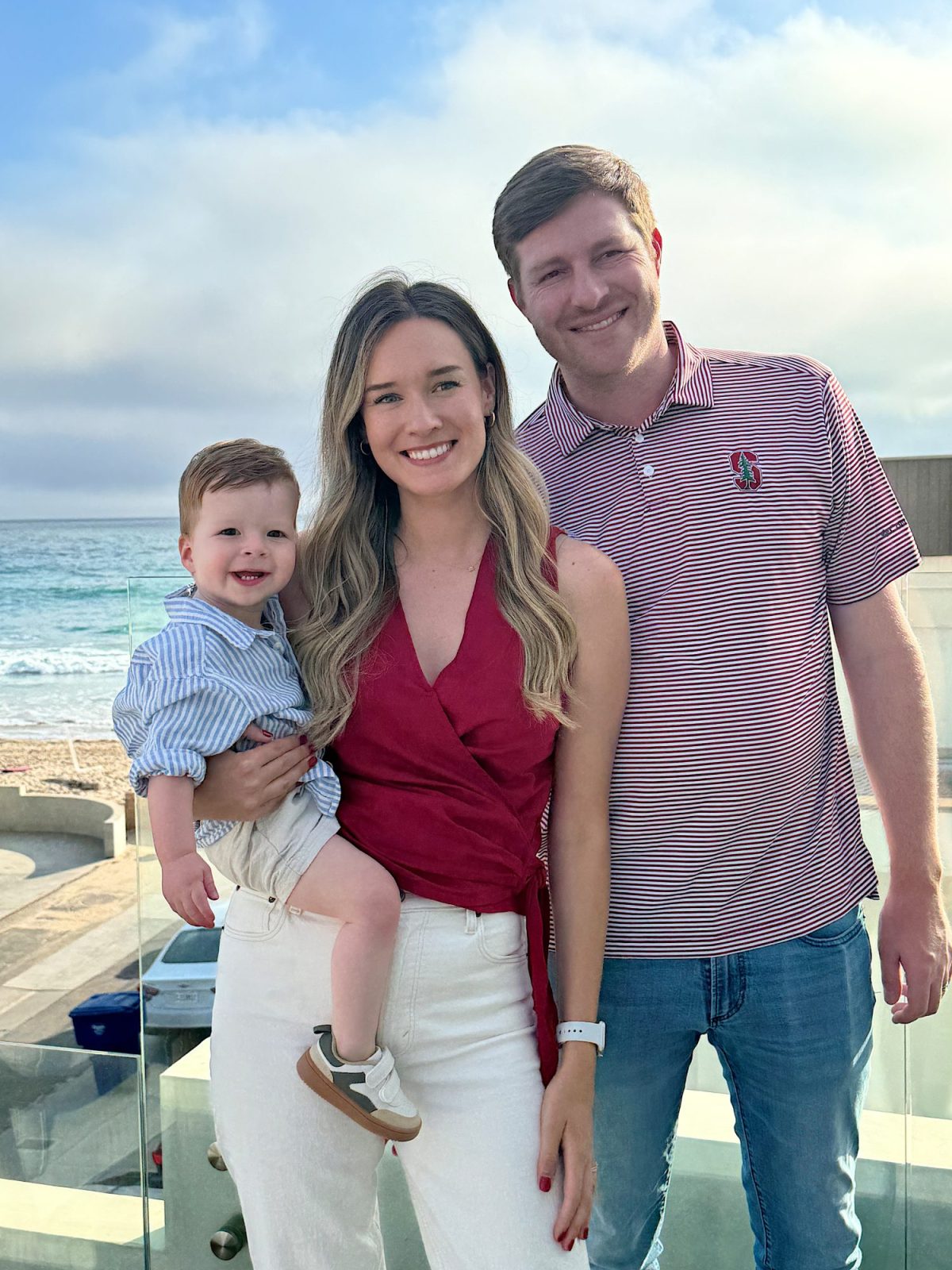 A woman holding a smiling toddler and a man stand together on a balcony near the ocean, all smiling at the camera.