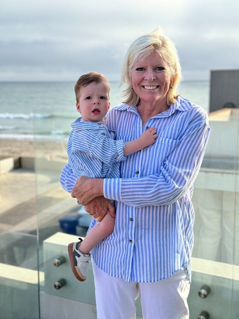 A woman smiles while holding a young child. They are both wearing light blue and white striped tops and standing in front of a beach scene.