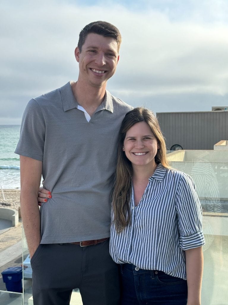 A man and woman are standing together on a seaside balcony, smiling at the camera. The man is wearing a grey polo shirt, and the woman is in a striped shirt. The ocean is visible in the background.