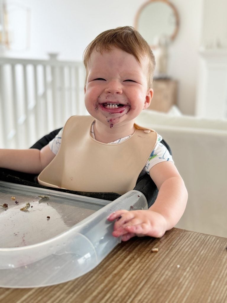 A young child with messy face and hands, wearing a bib, smiles widely while sitting in a high chair in a bright, modern room. Food remnants are on the tray and table.