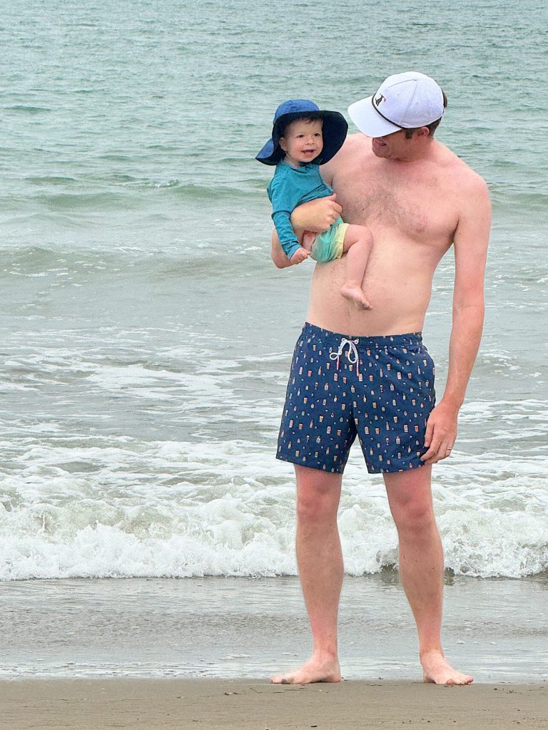 An adult man wearing a white cap and blue swim trunks holds a baby at the beach. The baby is in a blue sun hat and green swim diaper. They stand near the shoreline with waves in the background.