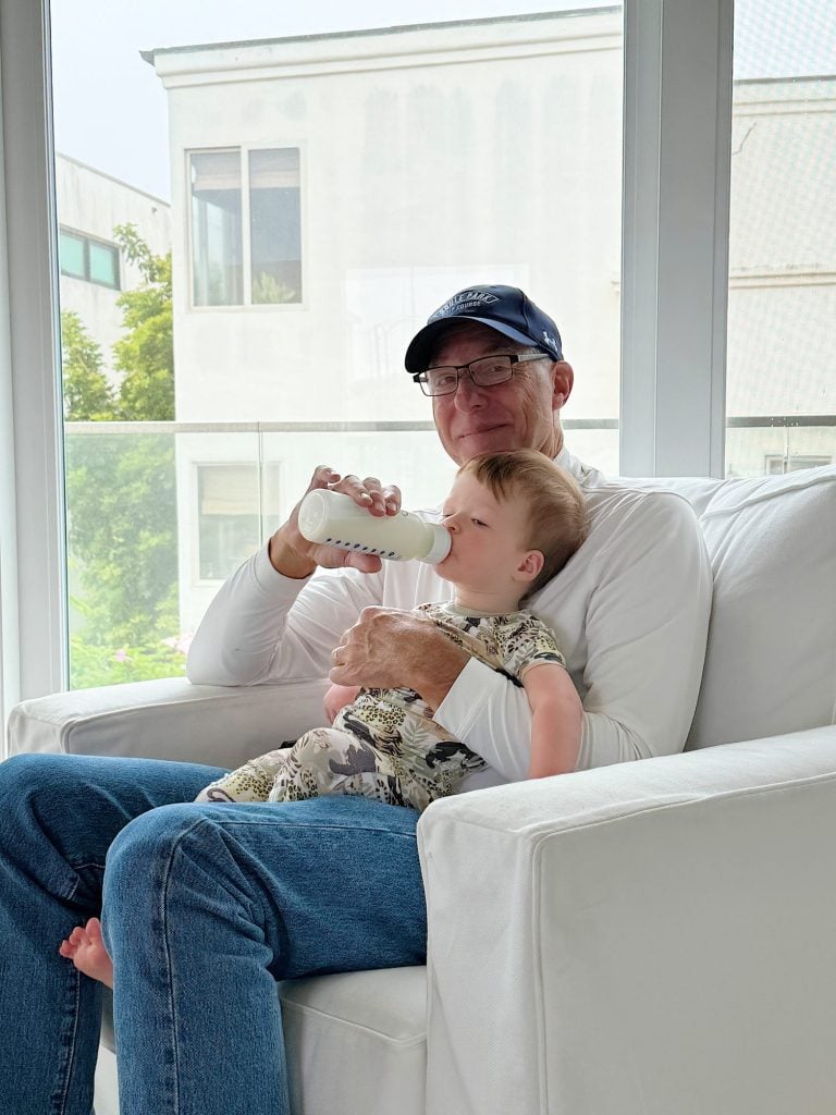 An older man wearing a cap is sitting on a white couch feeding milk from a bottle to a young child in his lap. They are in a bright room with large windows.