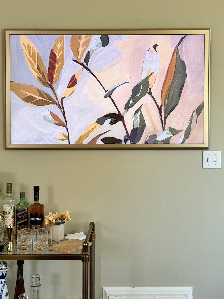 A framed abstract painting of leaves graces a beige wall above a bar cart laden with bottles, glasses, and a small flower arrangement—perfect for sprucing up your home decor in time for Amazon Prime Day 2023.