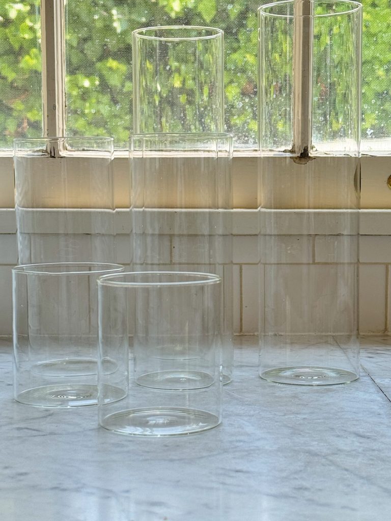 Five clear glass cylinders of varying heights are arranged on a marble countertop in front of a window with a garden view.