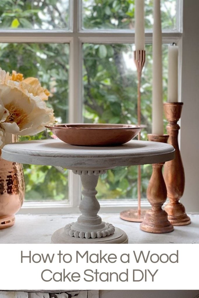 A wooden cake stand with a decorative base is placed on a windowsill with tall candles and flowers in the background. The text reads, "How to Make a Wood Cake Stand DIY.