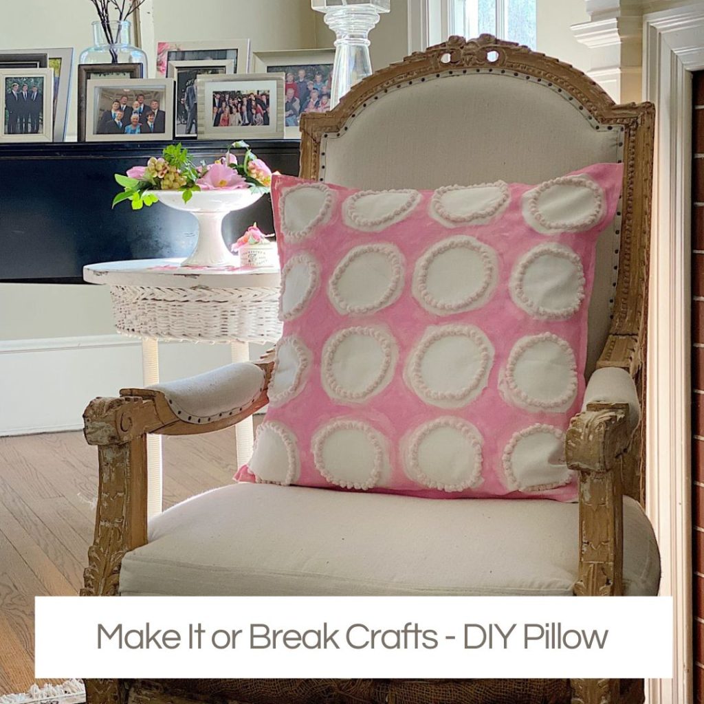A pink and white decorative pillow with a circular pattern, crafted with Creative Crafts flair, sits on a white upholstered chair in a well-lit room accented by framed photos in the background.