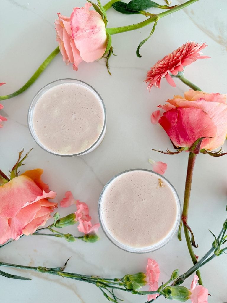 Two glasses of a creamy pink beverage surrounded by various pink flowers, including roses and carnations, on a white marbled surface.