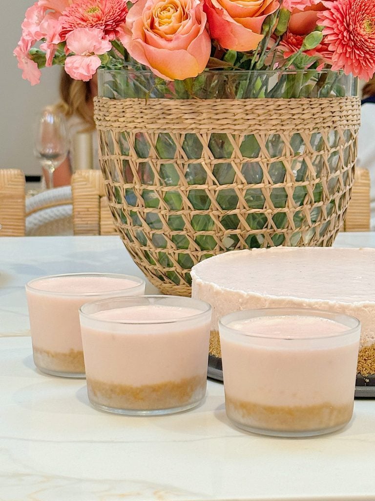 Three small glass cups and one larger round dish of cheesecake with pink topping next to a wicker basket with flowers.