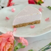 A slice of a pink cheesecake with a crumbly crust is on a white plate, surrounded by rose petals and pink flowers.