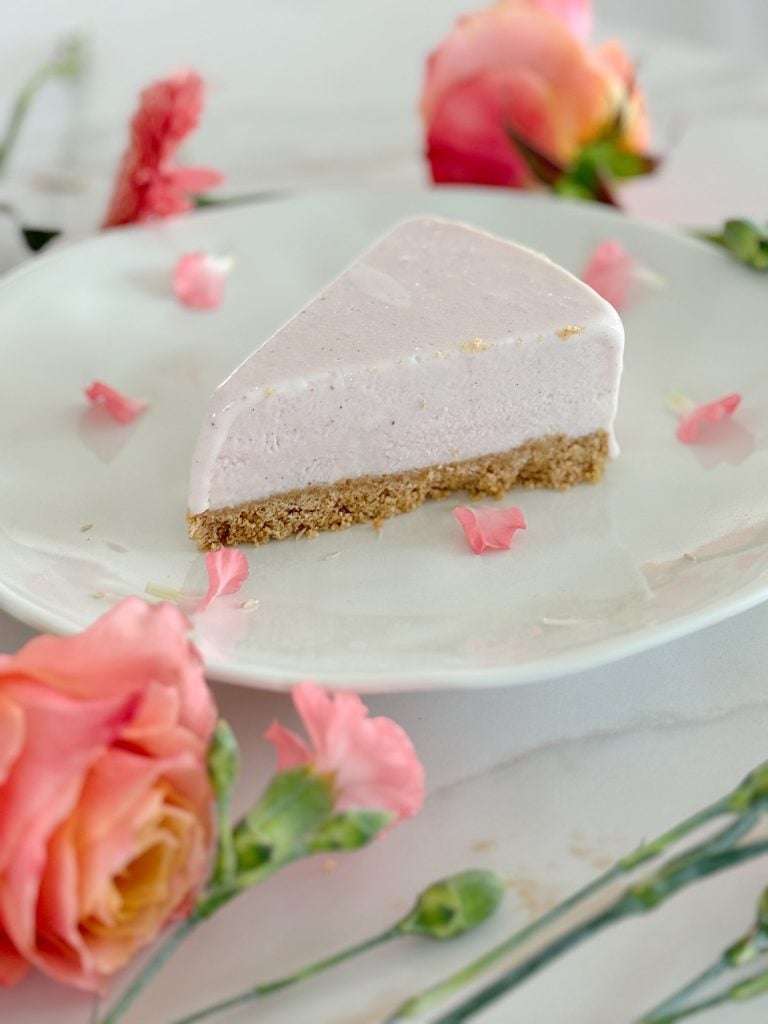 A slice of a pink cheesecake with a crumbly crust is on a white plate, surrounded by rose petals and pink flowers.