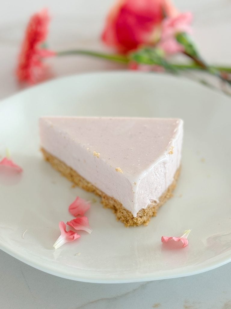 A slice of pink cheesecake with a graham cracker crust on a white plate, decorated with scattered pink flower petals.