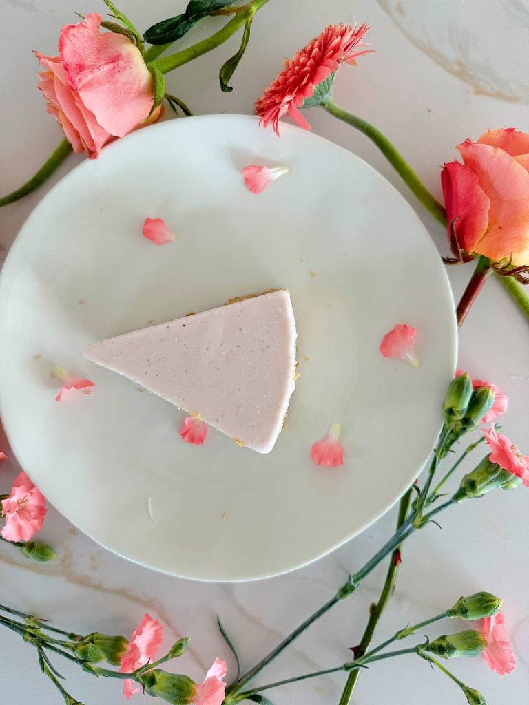 A slice of cake with white frosting on a white plate, surrounded by pink roses and petals on a marble surface.