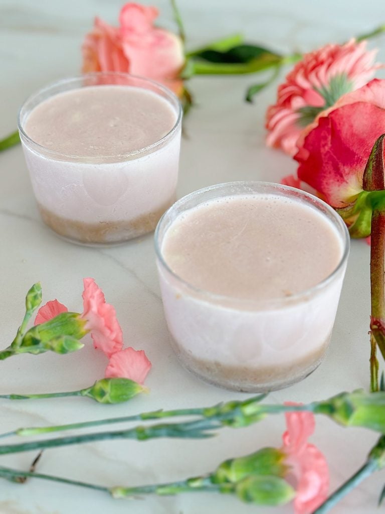 Two glasses with a pink beverage inside, placed on a white surface, surrounded by pink and red flowers.