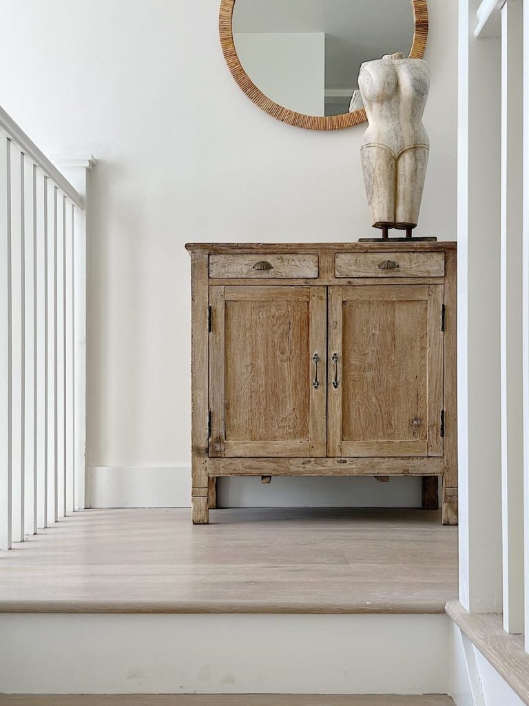 A rustic wooden cabinet with two doors is positioned at the top of a staircase, adorned with a torso statue and a round woven mirror hanging above it.