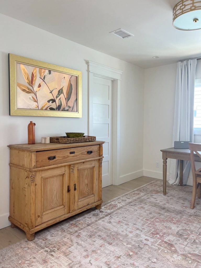 A light-filled room with a wooden sideboard, decorated with various items, below a framed botanical painting. A desk and chair sit beside a window with light curtains. A patterned rug covers the floor.