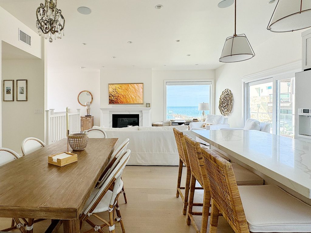 Modern open-concept living and dining area with white walls, wooden table and chairs, wicker bar stools, hanging pendant lights, and a fireplace with a TV above. Large windows offer natural light.