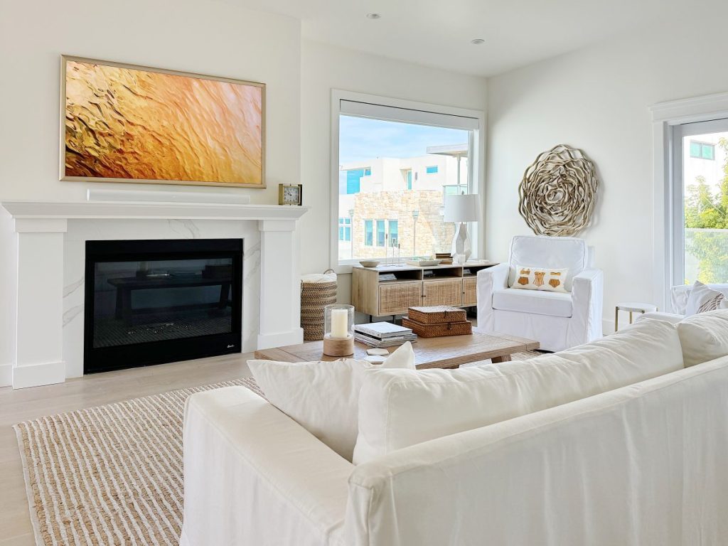 A modern living room features a white sofa, armchair, coffee table, and a fireplace with a golden abstract painting above it. Large windows provide natural light and a view outside.