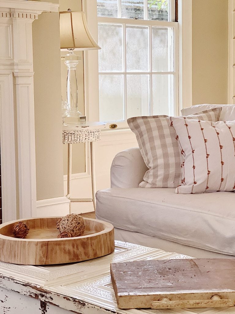 A cozy living room with a white sofa, striped and patterned pillows, a wooden bowl with decorative balls, and a white side table with a lamp next to a window.