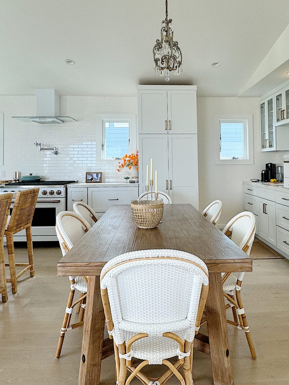 A modern kitchen features a wooden dining table with six wicker chairs, white cabinets, a stove with a hood, a chandelier, and a small bouquet of orange flowers on the counter.