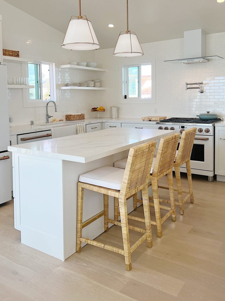 A modern kitchen with white cabinetry, a large island with three woven bar stools, pendant lights, open shelves with dishes, a stove with oven, and a window above the sink.