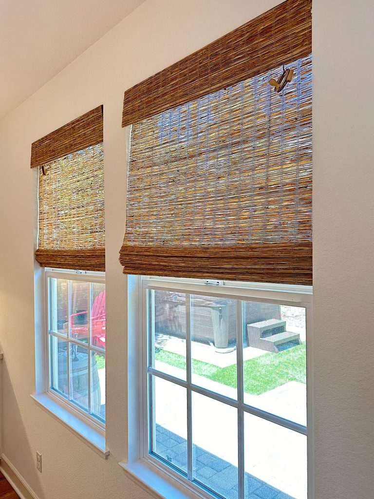 Two windows with bamboo blinds partially drawn, showing a view of a backyard with grass and a wooden deck.