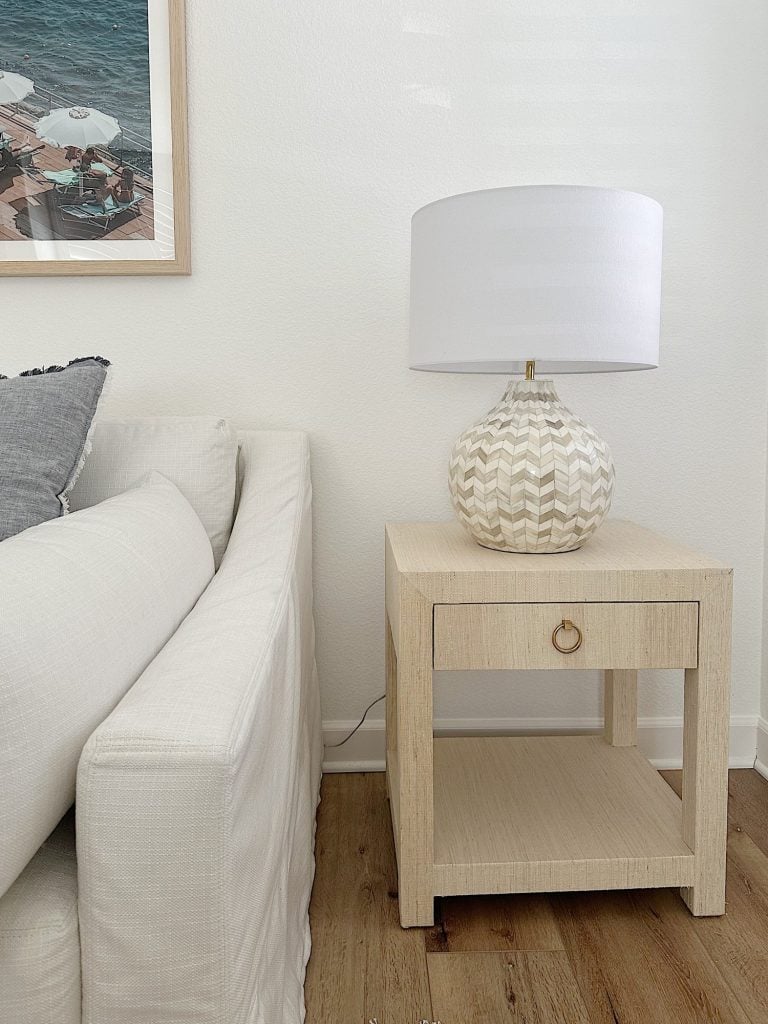 A light wooden side table with a drawer supports a patterned lamp with a white shade beside a white sofa. A framed photo hangs on the wall above. The floor is wooden.