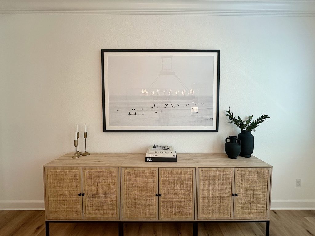 Sideboard with wicker doors, topped with two candle holders, a typewriter, and two large vases. Above it, a framed black-and-white photo is centered on a white wall.