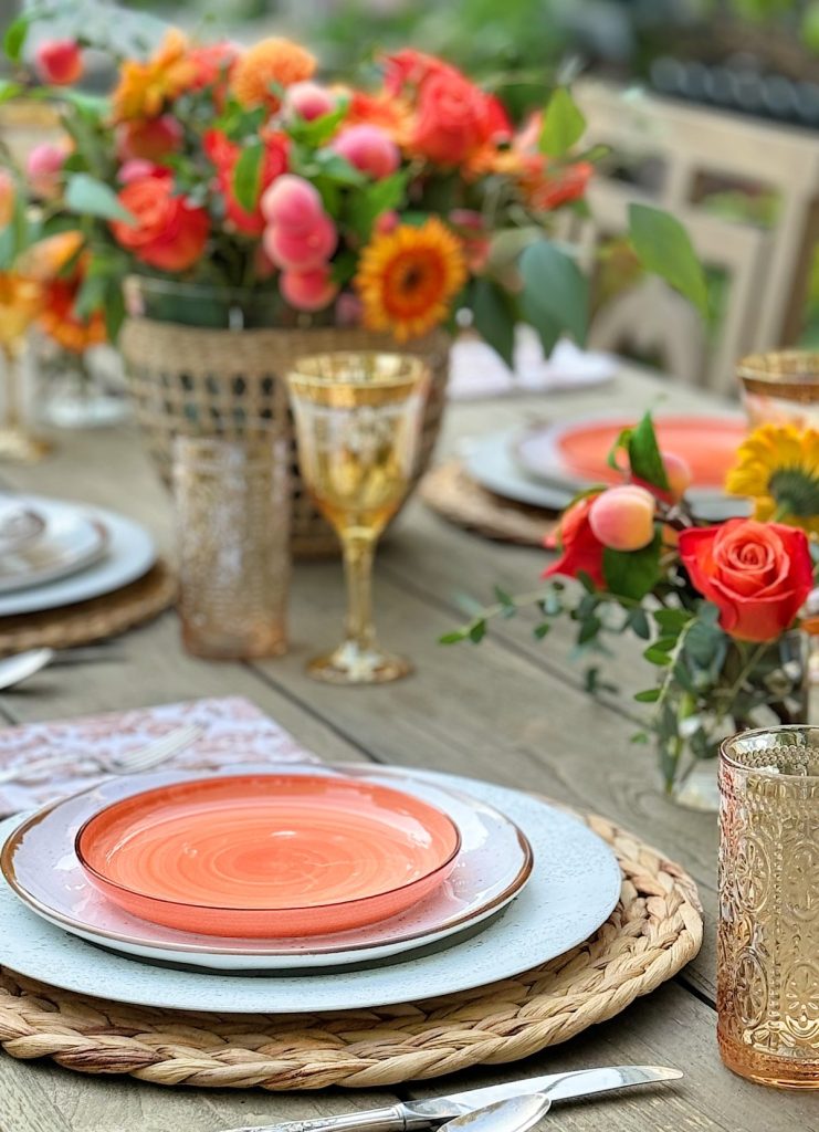 A dining table set with orange plates, golden glasses, woven placemats, and floral centerpieces featuring roses and orange flowers.