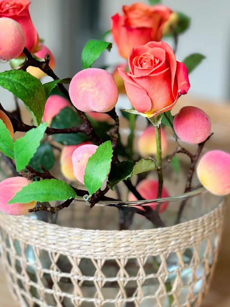 A clear glass vase with a woven rope covering holds artificial pink and peach flowers and leaves on a light wooden table.