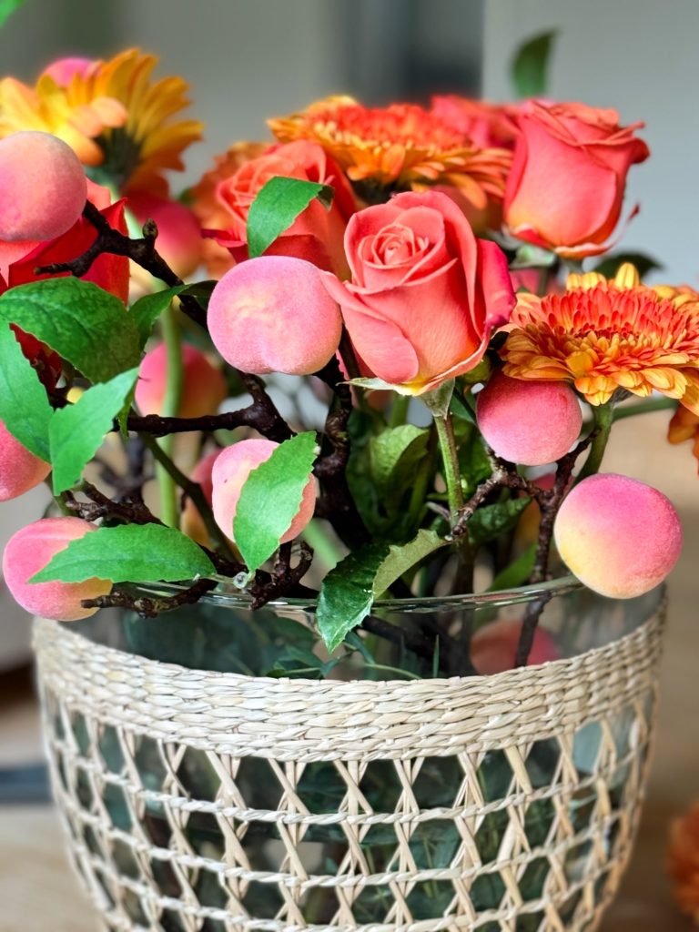 A woven basket holds a glass vase filled with pink roses, orange chrysanthemums, and peach branches with green leaves.