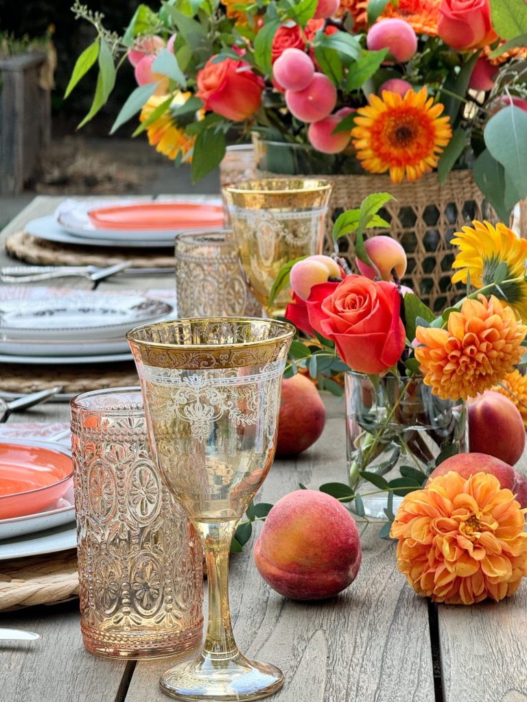 Outdoor table setting with ornate glassware, orange plates, and an arrangement of colorful flowers and peaches on a wooden table.