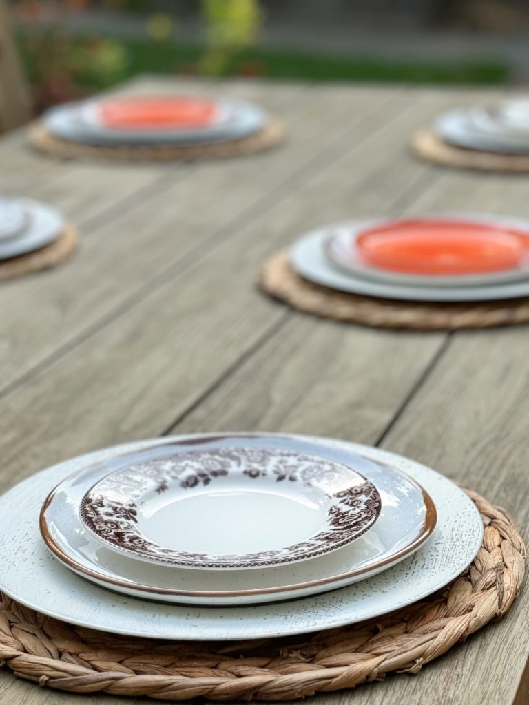 A wooden table is set with woven placemats and stacked china plates, including decorative ones with brown patterns and orange ones, arranged for a meal.