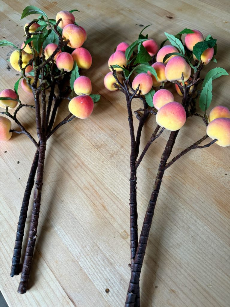 Two artificial peach branch decorations with multiple peaches and leaves lie on a wooden surface.