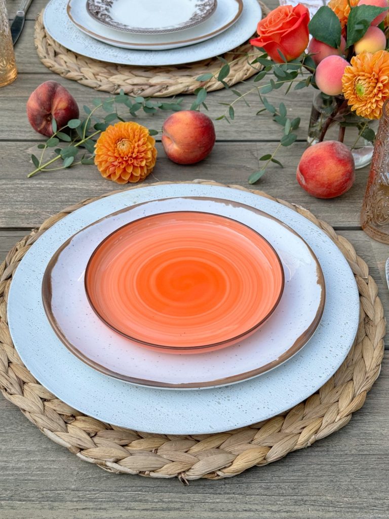 A table setting includes three stacked plates on a woven placemat. Flowers, peaches, and a floral arrangement decorate the table.