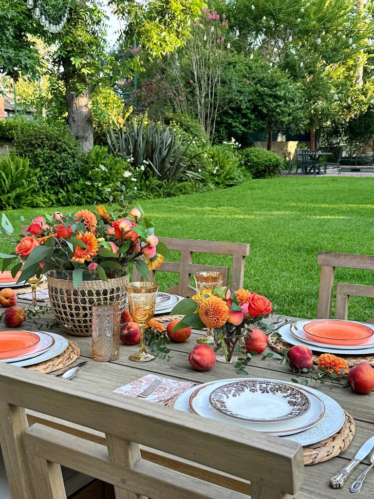 Outdoor dining table set with elegant place settings, flowers, peaches, and decorative plates, surrounded by a lush green garden.