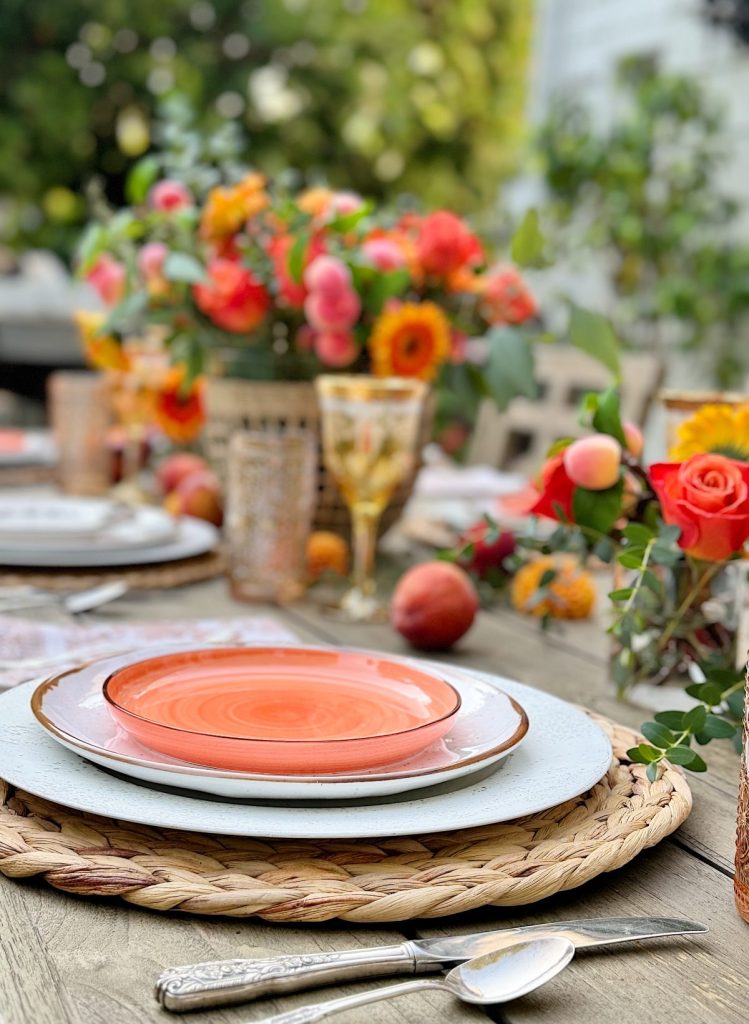 A table set for a meal with orange plates, woven placemats, silver cutlery, and floral centerpieces with vibrant orange and pink flowers. Peaches and glasses are also arranged on the wooden table.