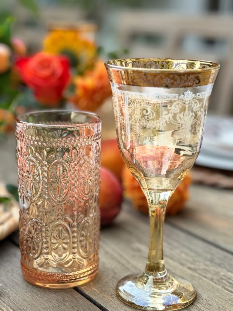 Two ornate glass goblets, one taller and one shorter, sit on a wooden table decorated with flowers in the background.