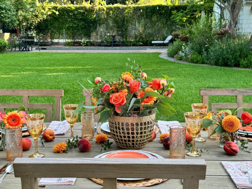 A wooden outdoor table is set with plates, glasses, peaches, and a floral centerpiece featuring orange and yellow flowers. The table is located in a lush, green garden.