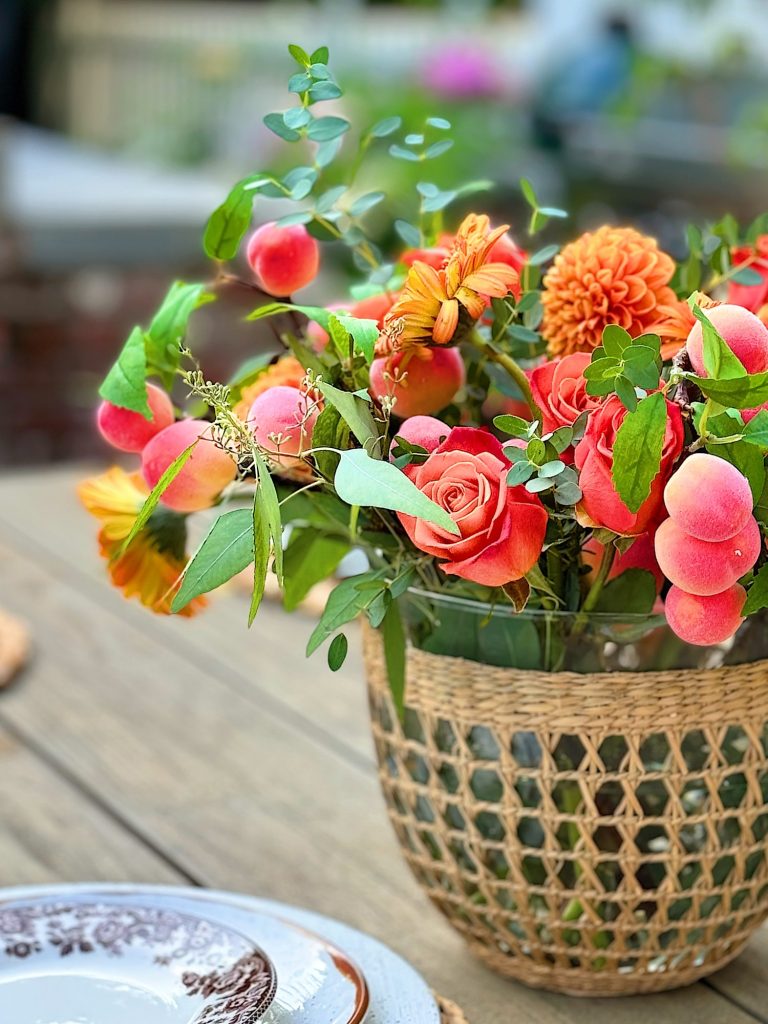 A wicker vase holds a vibrant bouquet of orange roses, dahlias, and greenery. The arrangement is placed on a wooden table next to patterned plates.