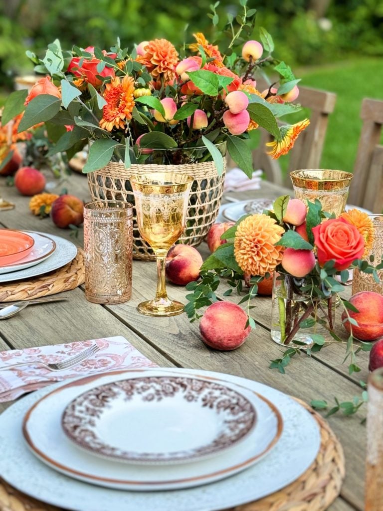A wooden table is set with plates, gold-rimmed glasses, and floral arrangements featuring orange and pink flowers. Peaches are scattered around the table as decorative elements.