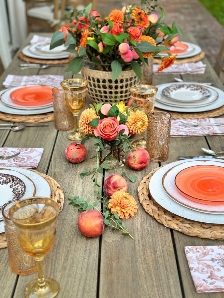 A wooden table is set for a meal with orange-themed plates, glasses, and napkins. A central floral arrangement includes roses and other orange flowers. Peaches are scattered as decoration.