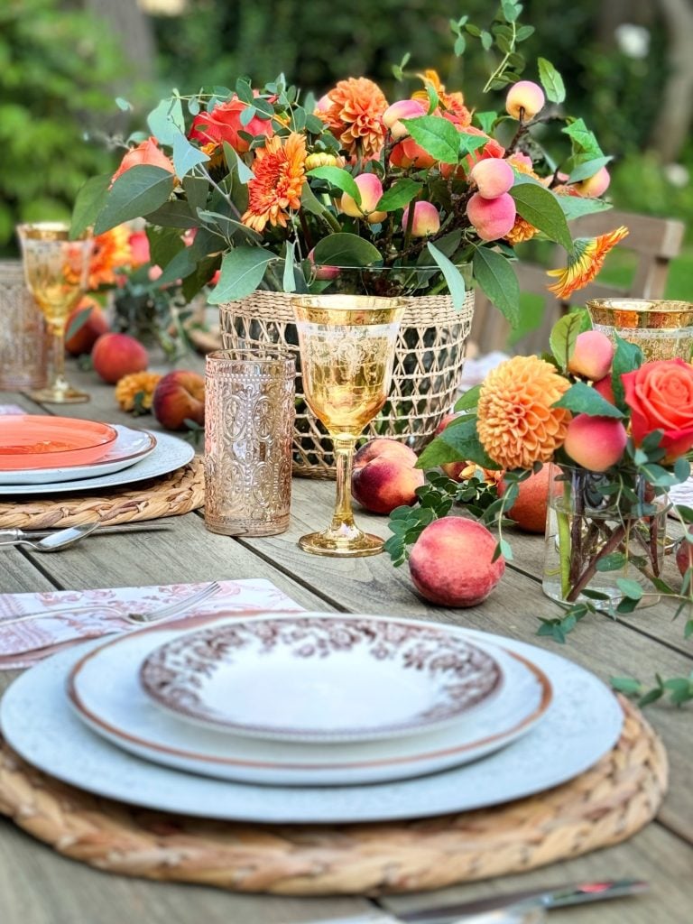 A rustic outdoor table setting with plates, cutlery, and glasses. The centerpiece includes orange flowers and peaches. There are additional fruits and orange roses beside the tableware.