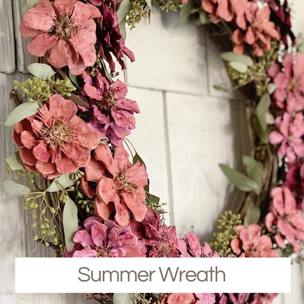 A decorative wreath with pink and red flowers and green foliage hangs on a light-colored wall. Text at the bottom reads "Summer Wreath." Perfect for Creative Crafts enthusiasts seeking a vibrant seasonal touch.