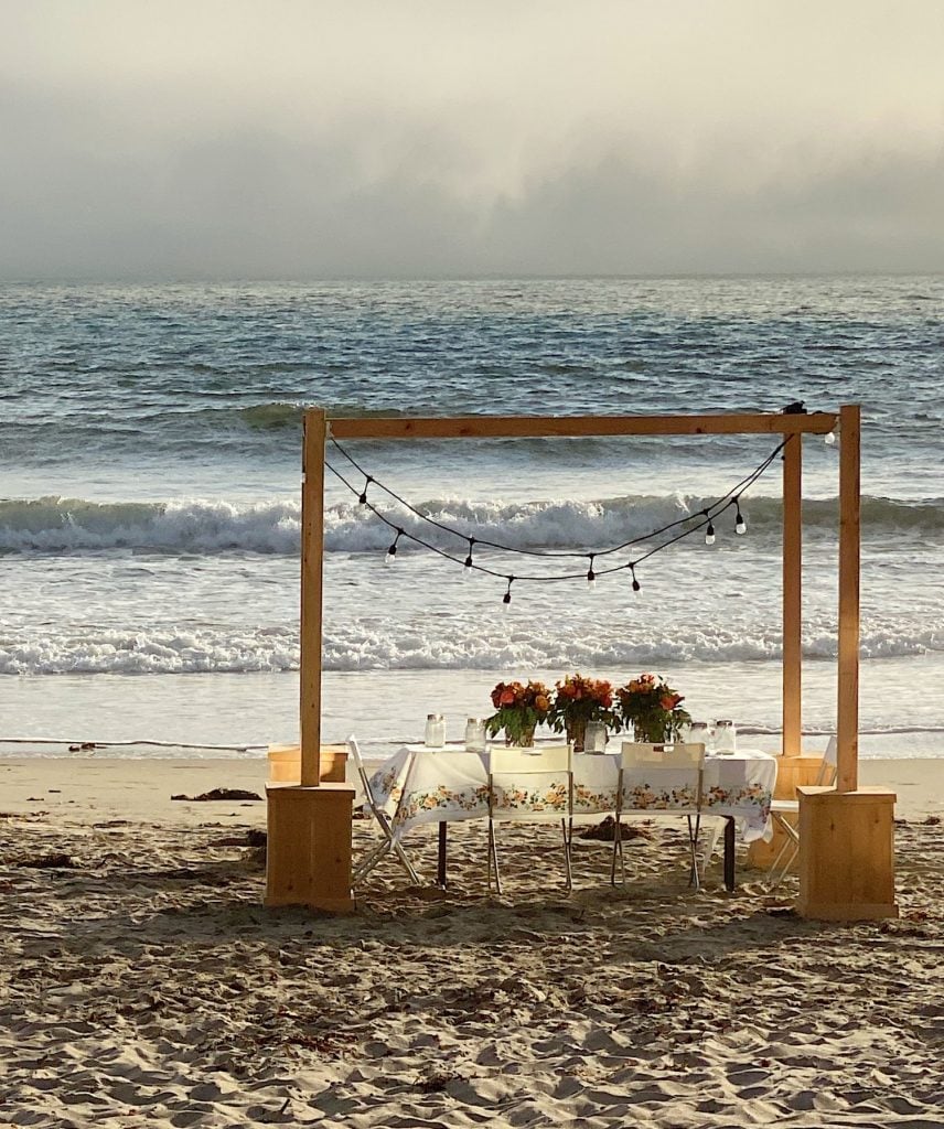 A dining table set up on a sandy beach under a wooden frame with string lights, facing the ocean. The table is adorned with flowers and tableware. Waves are visible in the background.